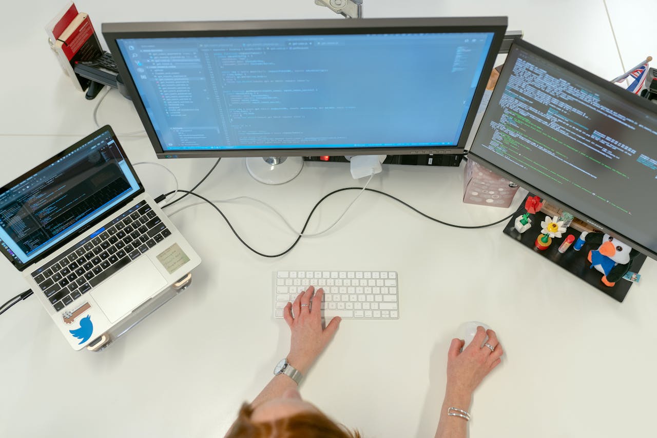 hero-img-01 A female software engineer coding on dual monitors and a laptop in an office setting.
